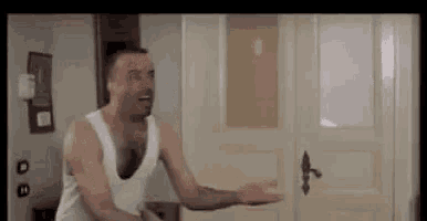 a man in a white tank top is standing in front of a door and laughing .