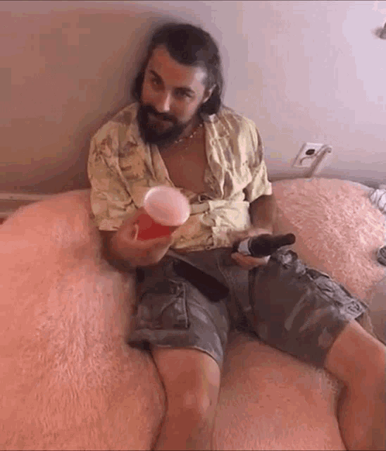 a man with a beard is sitting on a bed holding a beer and a cup