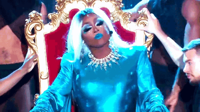 a drag queen in a blue dress is sitting on a red throne