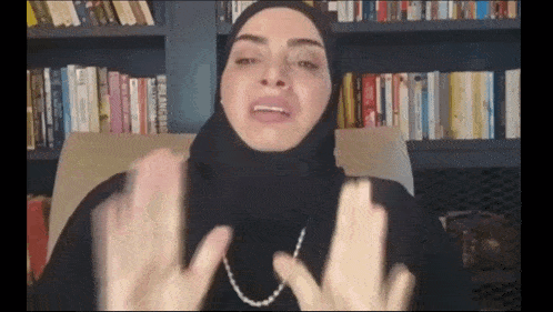 a woman wearing a hijab is sitting in front of a bookshelf with books on it .