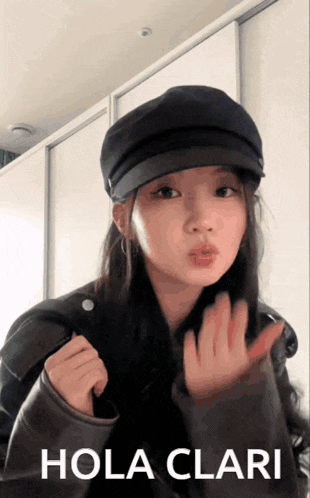 a woman wearing a black hat and a jacket says hola clari