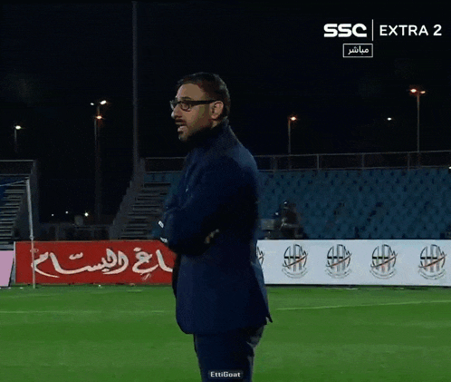 a man in a suit stands on a soccer field in front of a banner that says tameeni