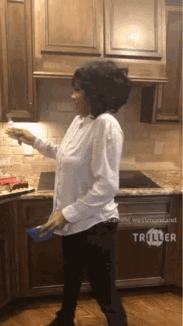 a woman in a white shirt is standing in a kitchen with triller on the bottom right