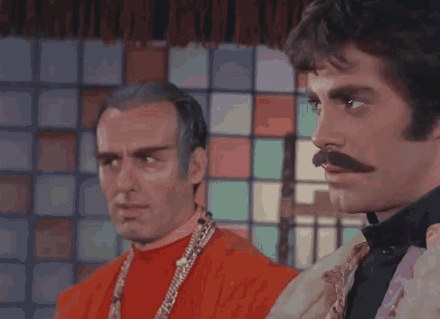 a man with a mustache is standing next to another man in an orange robe