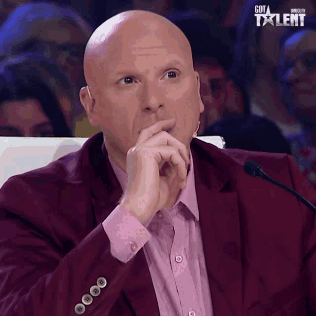 a bald man in a maroon suit and pink shirt is sitting in front of a microphone with got talent written on the screen behind him