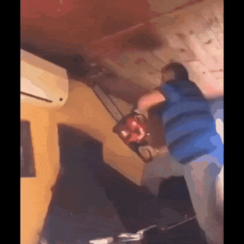 a man in a blue shirt is holding a chainsaw in a room with a ceiling fan .