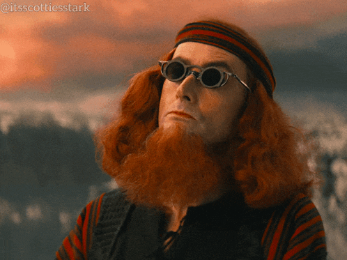a man with red hair and a beard wears sunglasses and a headband
