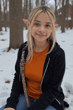 a woman wearing a black hoodie and an orange shirt is sitting in the snow holding a cane