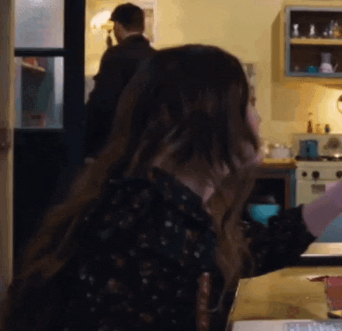 a woman is sitting at a table in a kitchen with a man standing in the background .