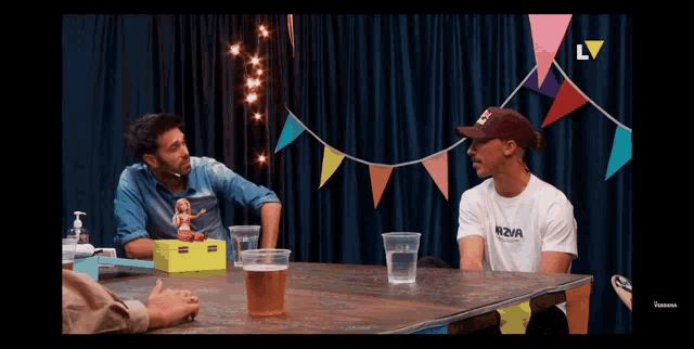 two men are sitting at a table and one has a shirt that says nova on it