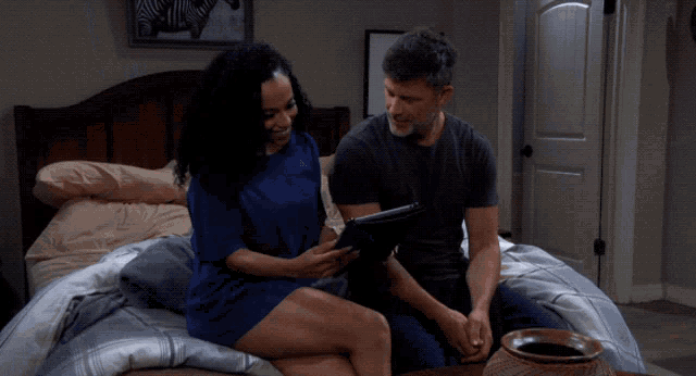 a man and woman are sitting on a bed looking at a tablet