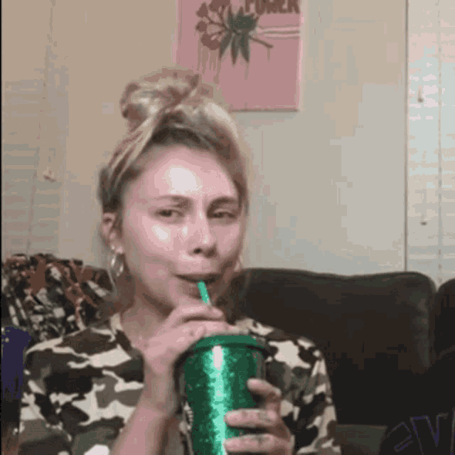 a woman in a camo shirt is drinking from a green cup with a straw .