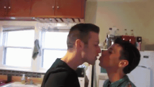two men are kissing in a kitchen and one of them is wearing a shirt that says ' coca cola ' on it