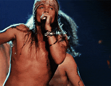a shirtless man singing into a microphone with a bandana around his head