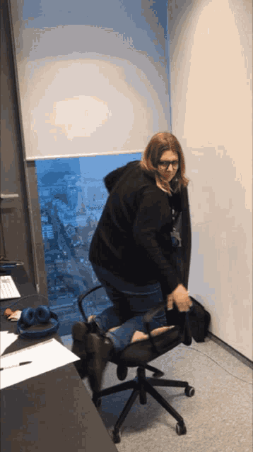 a woman in a black hoodie is kneeling down on an office chair in front of a window