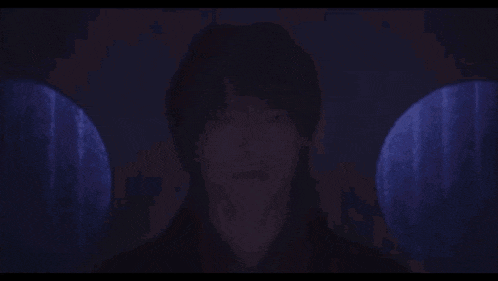 a man with long hair is standing in a dark room with a purple light behind him .