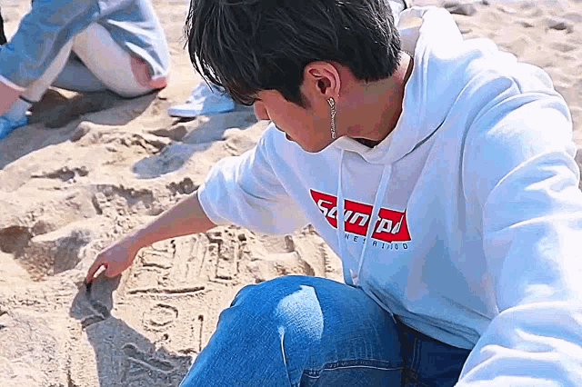 a man wearing a supreme hoodie is sitting on the beach drawing in the sand .