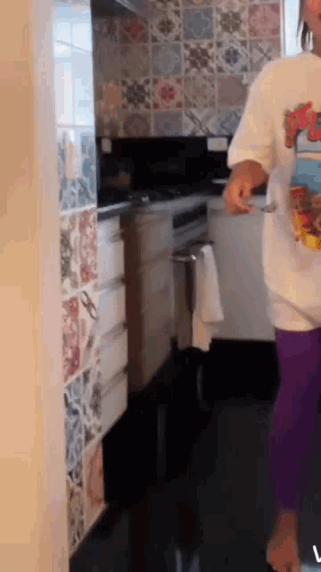 a woman in a white shirt and purple pants is standing in a kitchen holding a spoon .