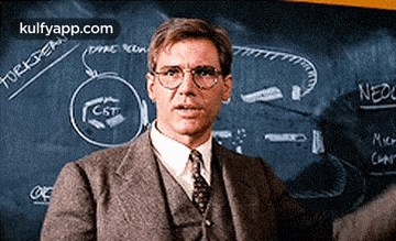 a man in a suit and tie is standing in front of a blackboard with a diagram on it .
