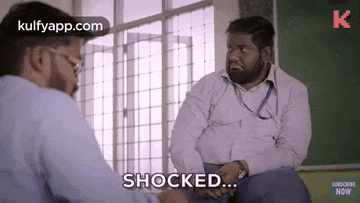 a man with a beard is sitting next to another man in a classroom and talking to him .