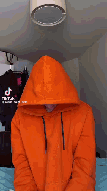 a man in an orange hoodie is making a funny face while sitting on a bed .