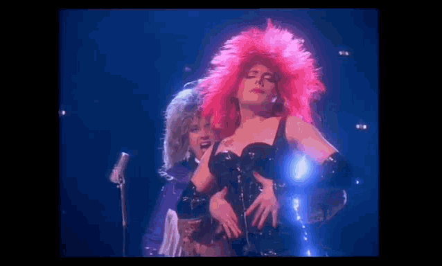 a woman in a pink wig is dancing on a stage next to a man in a purple dress .