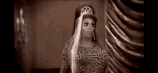 a drag queen wearing a leopard print dress and a tiara is standing in front of a wall .