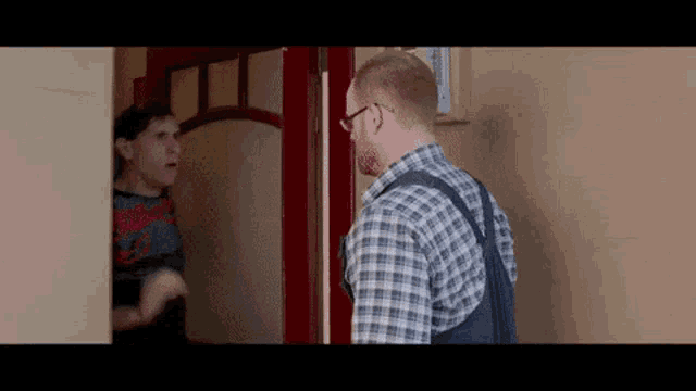 a man in a plaid shirt and overalls is standing in front of a door talking to another man .