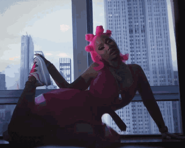 a woman with pink hair is kneeling down in front of a window with a city skyline in the background
