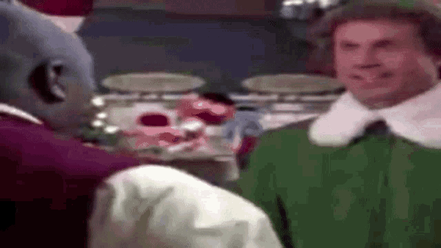 a man in a green elf costume is talking to a man in a red shirt .