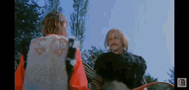a man in a fur vest talks to another man in a red shirt