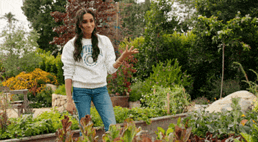 a woman in a white sweatshirt is standing in a garden surrounded by plants and flowers .