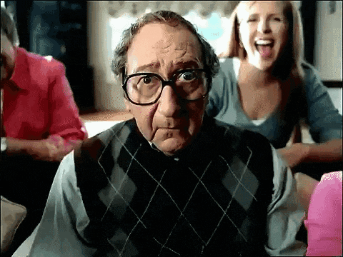 an elderly man wearing glasses and a plaid vest looks at the camera while a woman laughs behind him .