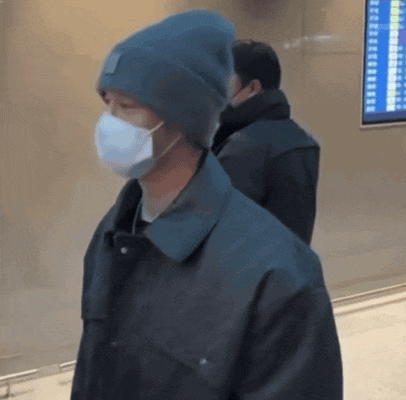 a man wearing a mask and a beanie stands in front of a screen that says ' departures ' on it