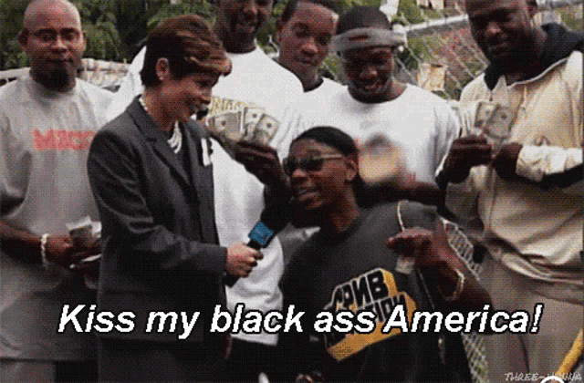a man wearing a shirt that says " kiss my black ass america " is being interviewed by a woman in a suit