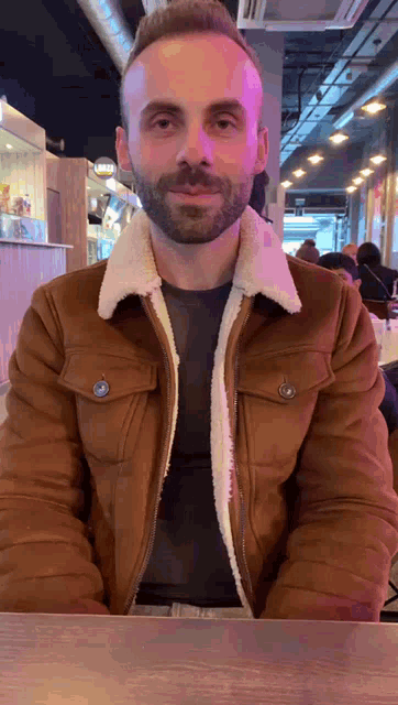 a man with a beard wearing a brown jacket is sitting at a table