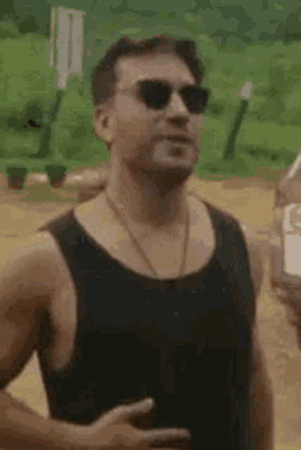 a man wearing sunglasses and a black tank top is smiling while standing next to a car .