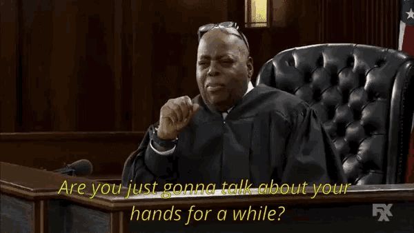 a man in a judge 's robe is sitting in a courtroom and talking about his hands for a while