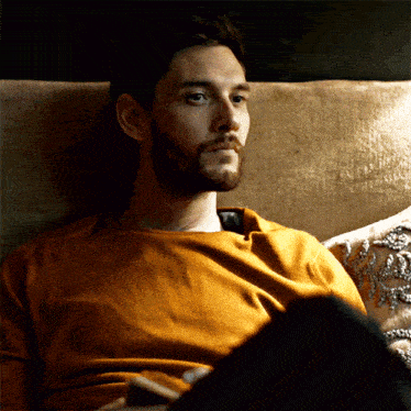 a man with a beard sits on a couch wearing a yellow shirt