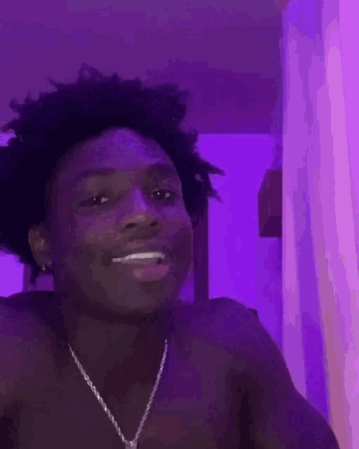 a shirtless young man wearing a necklace is smiling in front of a purple wall .