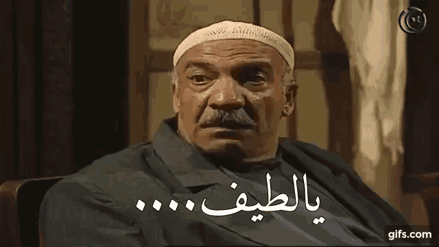 a man with a mustache and a hat is sitting in a chair with arabic writing on the screen .