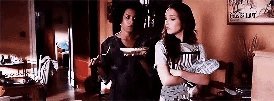 two women are standing next to each other in a living room . one of the women is holding a pie .