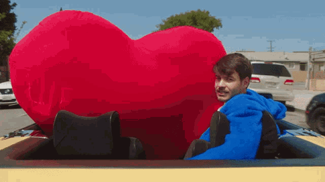 a man in a blue jacket is sitting in a car next to a large red heart shaped pillow
