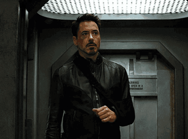 a man in a black leather jacket stands in front of a door that says warning