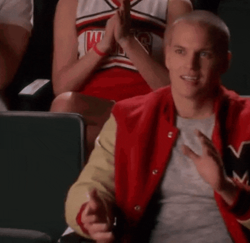 a man in a red and gold varsity jacket is sitting in a stadium clapping .