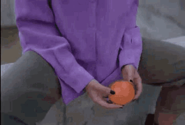a woman in a purple shirt is holding an orange .