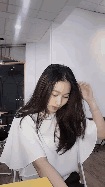a woman in a white top sits at a table with her hand in her hair