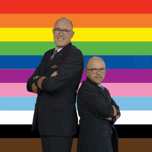 two men in suits standing back to back with their arms crossed in front of a rainbow flag