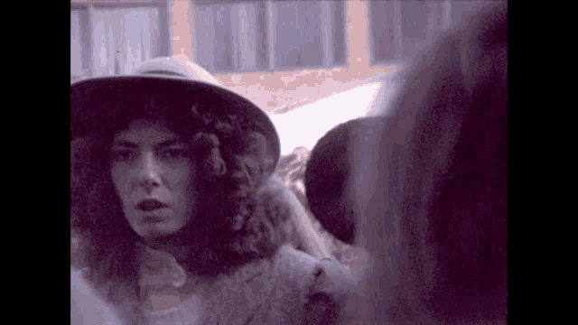 a woman with curly hair wearing a hat stands in front of a brick building