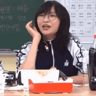 a woman wearing glasses is sitting at a table with a bottle of coke and a box of tissues